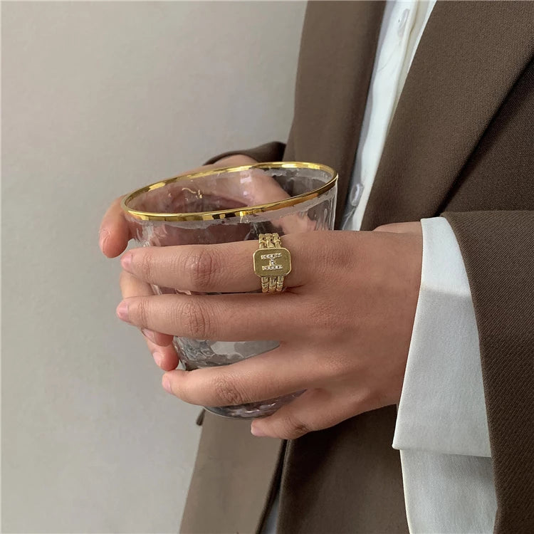 Gold ring with a brick-link band and a squared front plaque carrying a letter set with small clear stones
